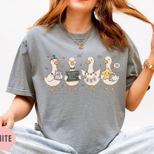 May include: Granite-colored t-shirt featuring a whimsical illustration of four cartoon geese. Each goose holds a different item, including a clipboard, a chalkboard with text, and a star. The shirt has a relaxed fit and short sleeves.