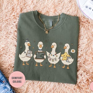 May include: Moss green t-shirt featuring a whimsical illustration of four cartoon ducks. Each duck is holding various items, including a chart, a sign with text, and a star. The shirt has a crew neck and short sleeves. A gold necklace is visible.