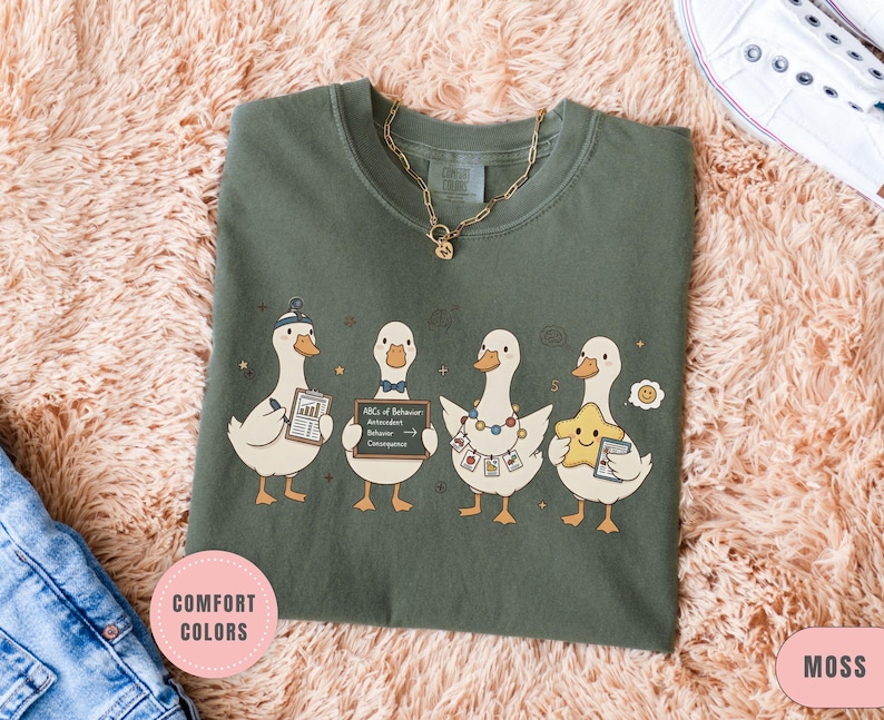 May include: Moss green t-shirt featuring a whimsical illustration of four cartoon ducks. Each duck is holding various items, including a chart, a sign with text, and a star. The shirt has a crew neck and short sleeves. A gold necklace is visible.