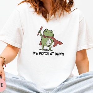 May include: A white t-shirt featuring a graphic of a green frog wearing a red cape, holding a red pen and a white book. The text below the frog reads "WE PSYCH AT DAWN" in black capital letters. The shirt has short sleeves and a crew neckline.