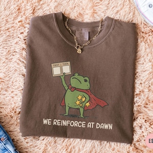 May include: Espresso-colored t-shirt featuring a cartoon frog superhero with a red cape and stars. The frog holds a sign that says "FIRST THEN" and the text "WE REINFORCE AT DAWN" is printed below the frog.