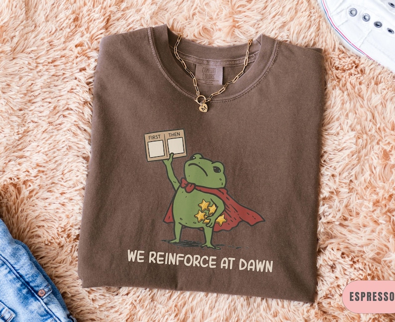 May include: Espresso-colored t-shirt featuring a cartoon frog superhero with a red cape and stars. The frog holds a sign that says "FIRST THEN" and the text "WE REINFORCE AT DAWN" is printed below the frog.