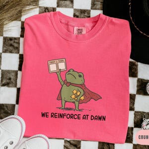 May include: A pink Comfort Colors sweatshirt featuring a cartoon frog superhero with a red cape and stars. The frog holds a sign that says "FIRST THEN". Below the frog, the text reads "WE REINFORCE AT DAWN".