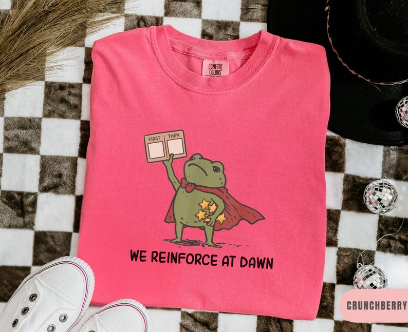 May include: A pink Comfort Colors sweatshirt featuring a cartoon frog superhero with a red cape and stars. The frog holds a sign that says "FIRST THEN". Below the frog, the text reads "WE REINFORCE AT DAWN".