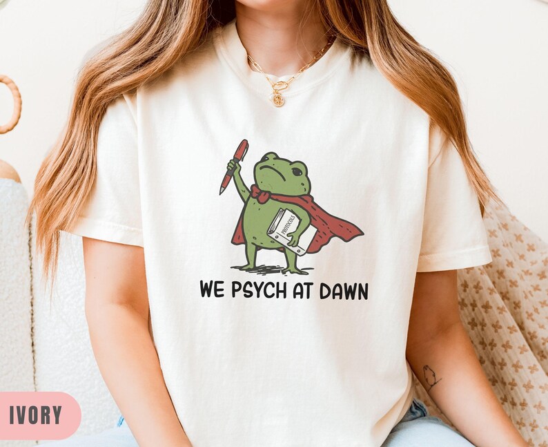 May include: An ivory t-shirt featuring a graphic of a green frog wearing a red cape and bow tie, holding a red pen and a white book. The text below the frog reads WE PSYCH AT DAWN in black capital letters. The shirt has short sleeves and a crew neck.