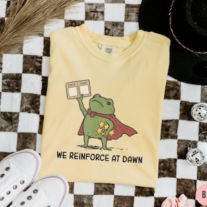 May include: Butter-colored t-shirt featuring a cartoon frog superhero with a red cape and holding a sign that says "First Then". The shirt also has the text "We Reinforce at Dawn".