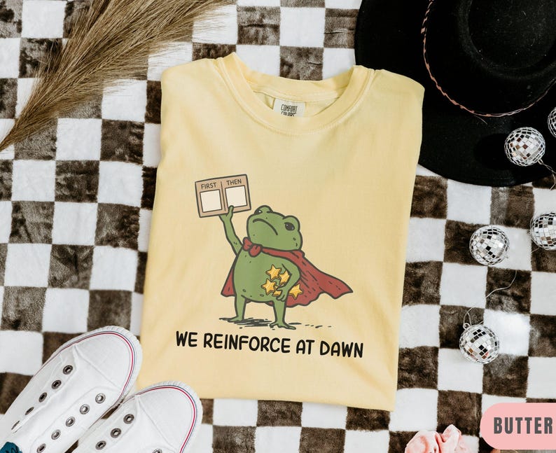 May include: Butter-colored t-shirt featuring a cartoon frog superhero with a red cape and holding a sign that says "First Then". The shirt also has the text "We Reinforce at Dawn".