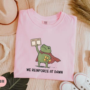 May include: A light pink t-shirt with a cartoon frog superhero graphic. The frog is wearing a cape and holding a sign that says "FIRST THEN". The text "WE REINFORCE AT DAWN" is printed below the frog. White sneakers are at the bottom.