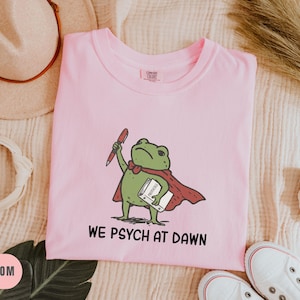 May include: A light pink t-shirt featuring a green frog illustration wearing a red cape, holding a red pen and a white book labeled Protocols. Below the graphic, the text reads WE PSYCH AT DAWN in black capital letters. The shirt has a crew neckline.