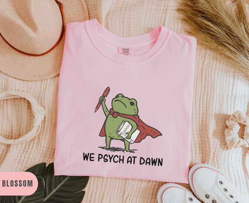 May include: A light pink t-shirt featuring a green frog illustration wearing a red cape, holding a red pen and a white book labeled Protocols. Below the graphic, the text reads WE PSYCH AT DAWN in black capital letters. The shirt has a crew neckline.