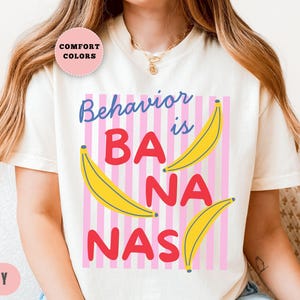 May include: Ivory-colored t-shirt with the phrase "Behavior is BANANAS" in blue and red letters, with yellow banana illustrations. The design is set against a pink and white striped background. The shirt has a round neckline and short sleeves.