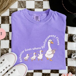 May include: Violet-colored t-shirt with a graphic of a goose wearing glasses and holding a notepad, leading a line of ducklings. The text "Behavior Goes where Reinforcement Flows" is printed on the shirt. White sneakers are visible in the bottom left corner.