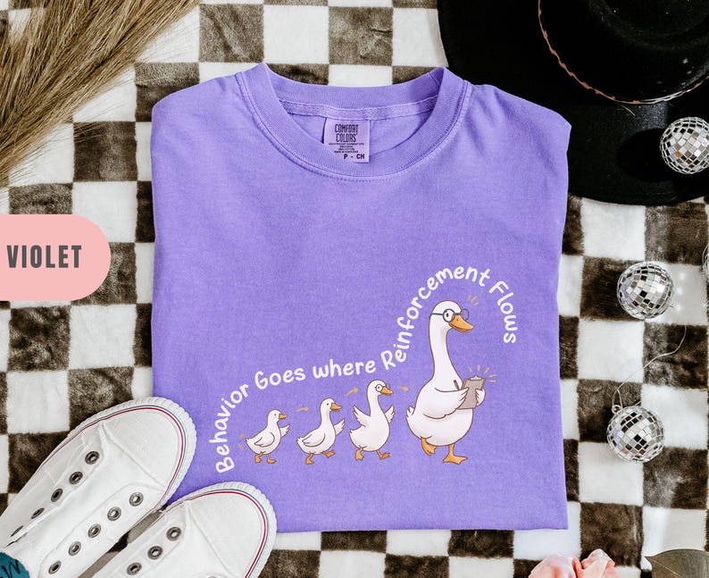 May include: Violet-colored t-shirt with a graphic of a goose wearing glasses and holding a notepad, leading a line of ducklings. The text "Behavior Goes where Reinforcement Flows" is printed on the shirt. White sneakers are visible in the bottom left corner.