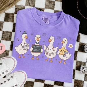 May include: A violet Comfort Colors t-shirt featuring a whimsical design of four cartoon geese. Each goose holds an item, including a clipboard, a sign reading "ABCs of Behavior," and a star. The shirt is laid flat on a checkered surface.