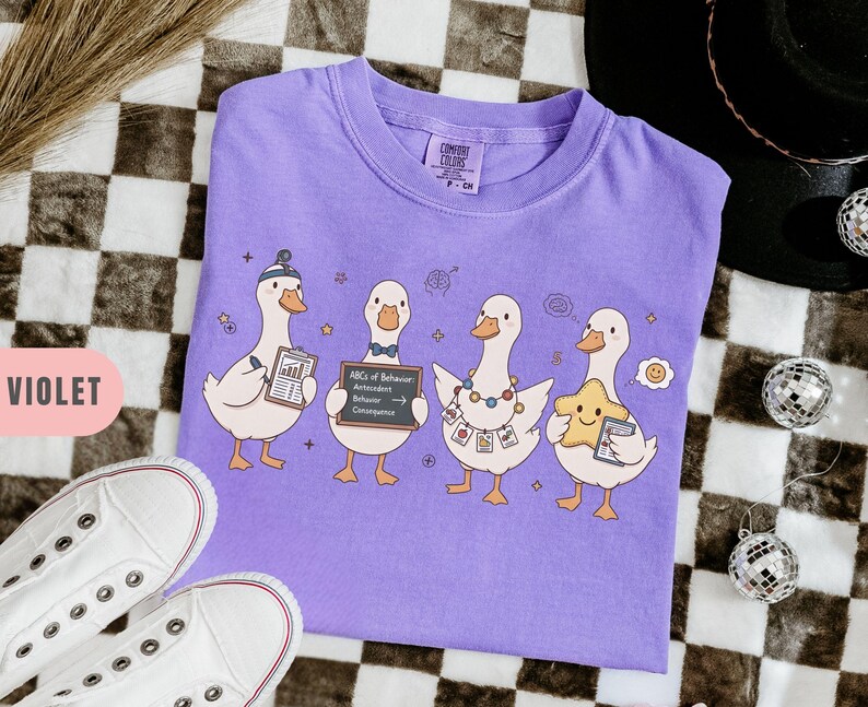 May include: A violet Comfort Colors t-shirt featuring a whimsical design of four cartoon geese. Each goose holds an item, including a clipboard, a sign reading "ABCs of Behavior," and a star. The shirt is laid flat on a checkered surface.