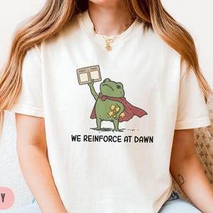 May include: Ivory-colored t-shirt featuring a cartoon frog superhero with a red cape and gold stars. The frog holds a sign that says "First Then". The text "WE REINFORCE AT DAWN" is printed below the frog.