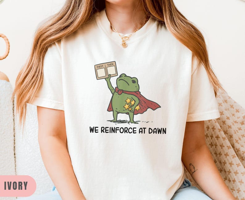 May include: Ivory-colored t-shirt featuring a cartoon frog superhero with a red cape and gold stars. The frog holds a sign that says "First Then". The text "WE REINFORCE AT DAWN" is printed below the frog.