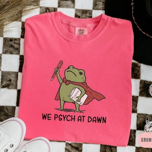 May include: A bright pink crewneck t-shirt featuring a graphic of a green frog wearing a red cape, holding a red pen and a book labeled Protocols. Below the frog, the text reads WE PSYCH AT DAWN in black capital letters. The shirt has a Comfort Colors tag.