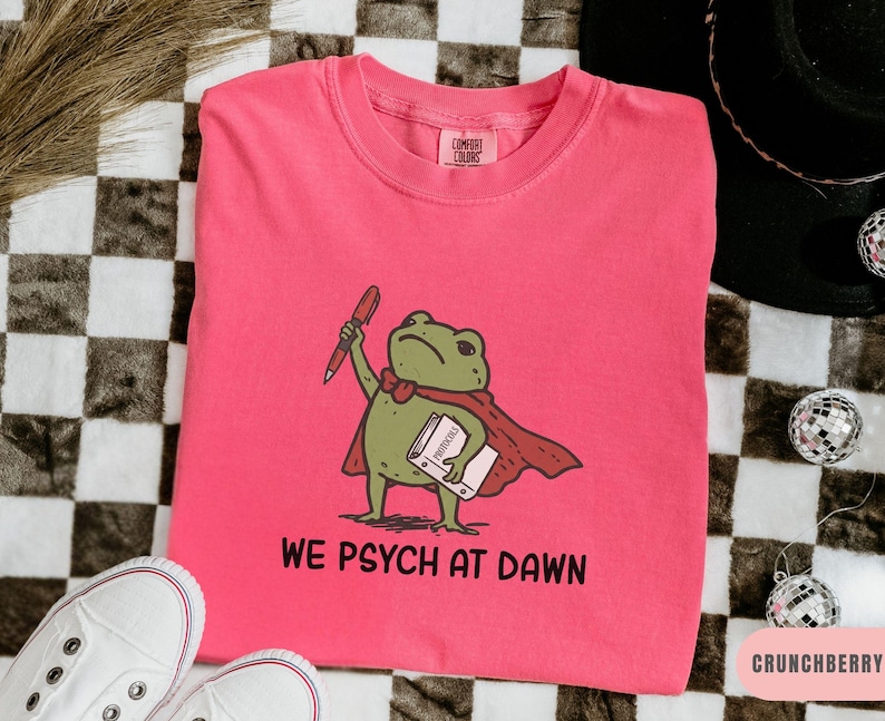 May include: A bright pink crewneck t-shirt featuring a graphic of a green frog wearing a red cape, holding a red pen and a book labeled Protocols. Below the frog, the text reads WE PSYCH AT DAWN in black capital letters. The shirt has a Comfort Colors tag.