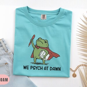 May include: A sea foam green t-shirt featuring a graphic of a green frog wearing a red cape, holding a red pen and a white book labeled Protocols. Below the frog, the text reads WE PSYCH AT DAWN in black capital letters. The shirt has a crew neck.