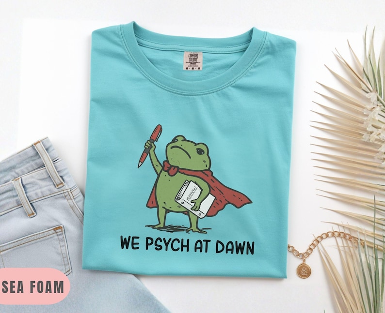 May include: A sea foam green t-shirt featuring a graphic of a green frog wearing a red cape, holding a red pen and a white book labeled Protocols. Below the frog, the text reads WE PSYCH AT DAWN in black capital letters. The shirt has a crew neck.