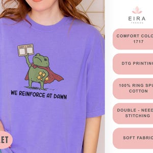 May include: A violet t-shirt features a cartoon frog superhero with a red cape and star-shaped badges. The frog holds a sign that says "FIRST THEN". The text "WE REINFORCE AT DAWN" is printed below the frog. The shirt is made of soft fabric.
