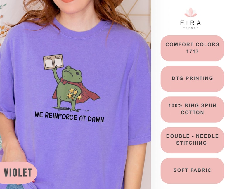 May include: A violet t-shirt features a cartoon frog superhero with a red cape and star-shaped badges. The frog holds a sign that says "FIRST THEN". The text "WE REINFORCE AT DAWN" is printed below the frog. The shirt is made of soft fabric.