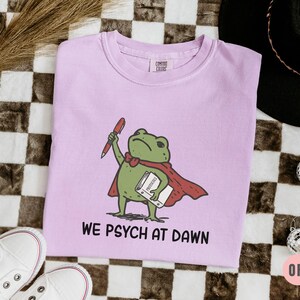 May include: A light purple t-shirt featuring a graphic of a green frog wearing a red cape, holding a red pen and a white book labeled "PROTOCOLS". Below the frog, the text "WE PSYCH AT DAWN" is printed in black capital letters. The shirt has a crew neck.