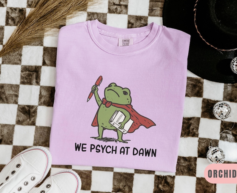 May include: A light purple t-shirt featuring a graphic of a green frog wearing a red cape, holding a red pen and a white book labeled "PROTOCOLS". Below the frog, the text "WE PSYCH AT DAWN" is printed in black capital letters. The shirt has a crew neck.
