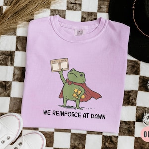 May include: A light purple t-shirt featuring a cartoon frog superhero with a red cape and holding a sign that says "First Then". The shirt also has the text "We Reinforce at Dawn". The shirt is next to white sneakers.