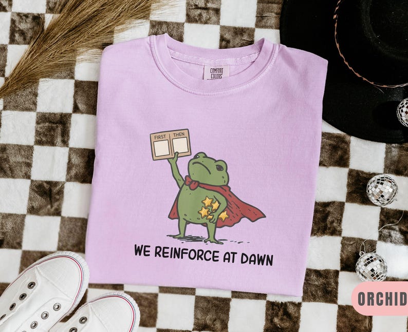 May include: A light purple t-shirt featuring a cartoon frog superhero with a red cape and holding a sign that says "First Then". The shirt also has the text "We Reinforce at Dawn". The shirt is next to white sneakers.