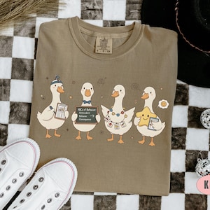 May include: Khaki-colored t-shirt featuring a whimsical illustration of four cartoon ducks. Each duck holds a different item, including a chart, a chalkboard with text, and a star. The shirt has a crew neck and short sleeves.