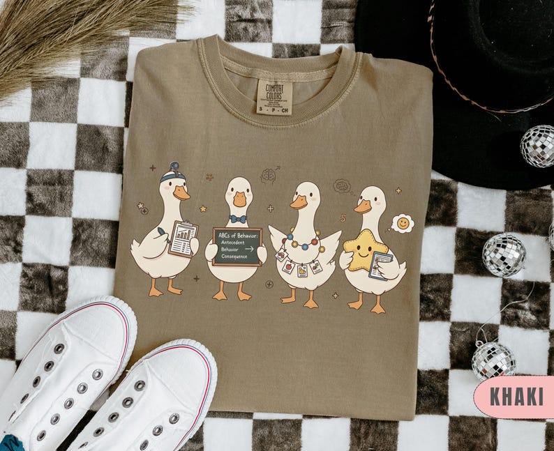 May include: Khaki-colored t-shirt featuring a whimsical illustration of four cartoon ducks. Each duck holds a different item, including a chart, a chalkboard with text, and a star. The shirt has a crew neck and short sleeves.