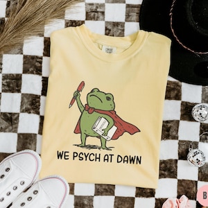 May include: A pale yellow short-sleeve t-shirt featuring a green frog wearing a red cape, holding a red pen and a white book labeled Protocols. Below the frog, the text reads WE PSYCH AT DAWN in black capital letters. The shirt has a crew neckline.