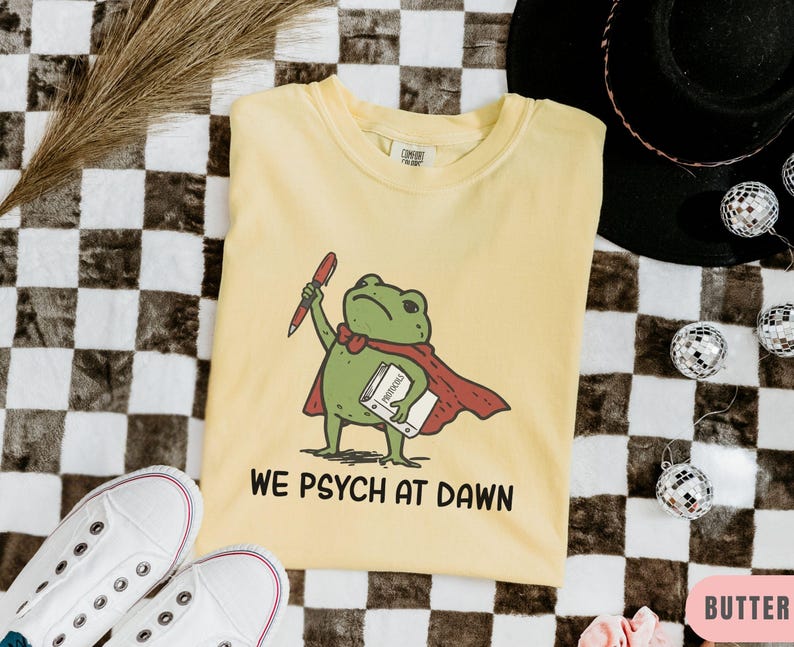 May include: A pale yellow short-sleeve t-shirt featuring a green frog wearing a red cape, holding a red pen and a white book labeled Protocols. Below the frog, the text reads WE PSYCH AT DAWN in black capital letters. The shirt has a crew neckline.