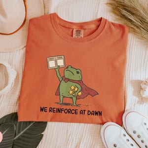 May include: Orange t-shirt featuring a cartoon frog superhero with a red cape and stars. The frog holds a sign that says "First Then". The text "WE REINFORCE AT DAWN" is printed below the frog. White sneakers are at the bottom.