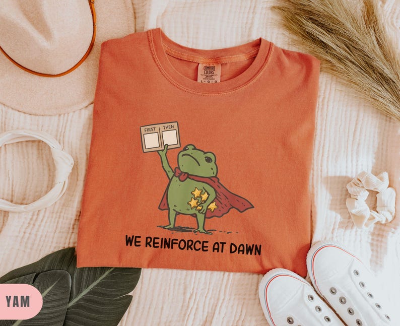 May include: Orange t-shirt featuring a cartoon frog superhero with a red cape and stars. The frog holds a sign that says "First Then". The text "WE REINFORCE AT DAWN" is printed below the frog. White sneakers are at the bottom.
