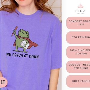 May include: A violet t-shirt featuring a graphic of a green frog wearing a red cape, holding a red pen and a white book. The text below the frog reads WE PSYCH AT DAWN. The shirt is made of 100% ring spun cotton with double-needle stitching.