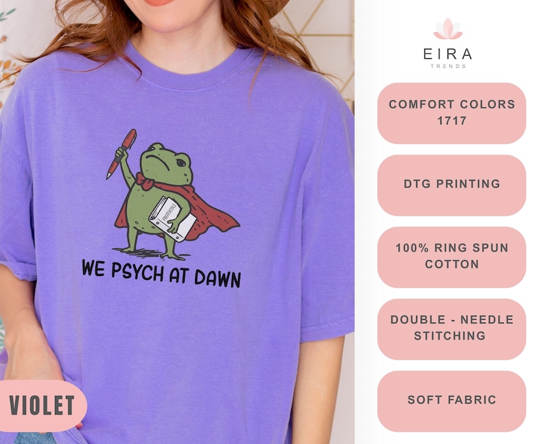 May include: A violet t-shirt featuring a graphic of a green frog wearing a red cape, holding a red pen and a white book. The text below the frog reads WE PSYCH AT DAWN. The shirt is made of 100% ring spun cotton with double-needle stitching.