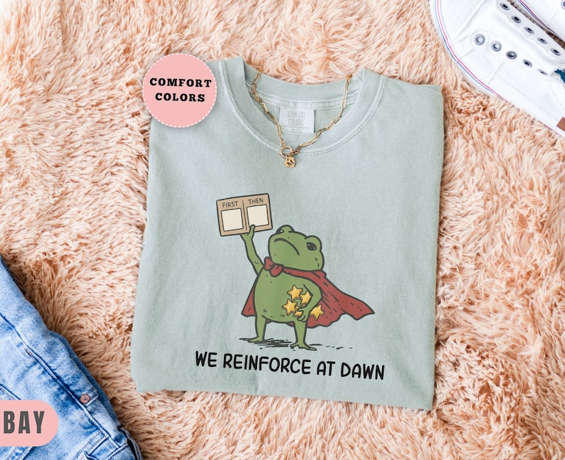 May include: A light green t-shirt featuring a cartoon frog superhero holding a "First Then" card. The frog wears a red cape and has gold stars. The text "WE REINFORCE AT DAWN" is printed below the frog.