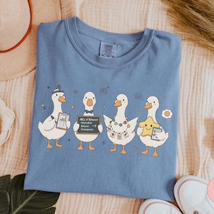 May include: Blue jean Comfort Colors t-shirt featuring four cartoon ducks. Each duck holds an item, including a chart, a chalkboard with text, and a star. The shirt is a solid blue color.