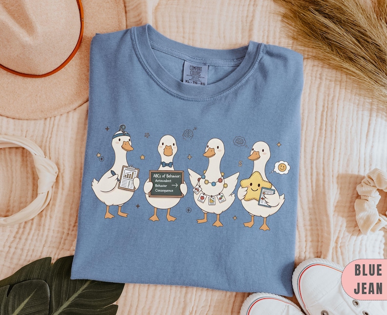 May include: Blue jean Comfort Colors t-shirt featuring four cartoon ducks. Each duck holds an item, including a chart, a chalkboard with text, and a star. The shirt is a solid blue color.