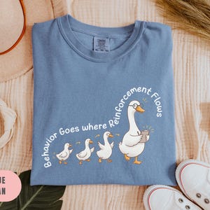 May include: Blue jean t-shirt with a crew neck. The shirt features a cartoon goose wearing glasses and holding a notepad, followed by three smaller geese. Text on the shirt reads "Behavior Goes where Reinforcement Flows."