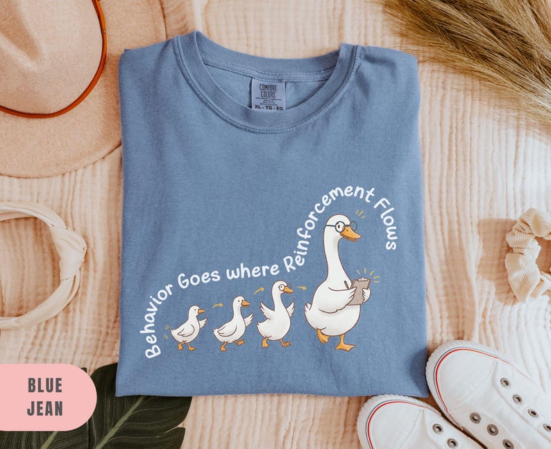 May include: Blue jean t-shirt with a crew neck. The shirt features a cartoon goose wearing glasses and holding a notepad, followed by three smaller geese. Text on the shirt reads "Behavior Goes where Reinforcement Flows."