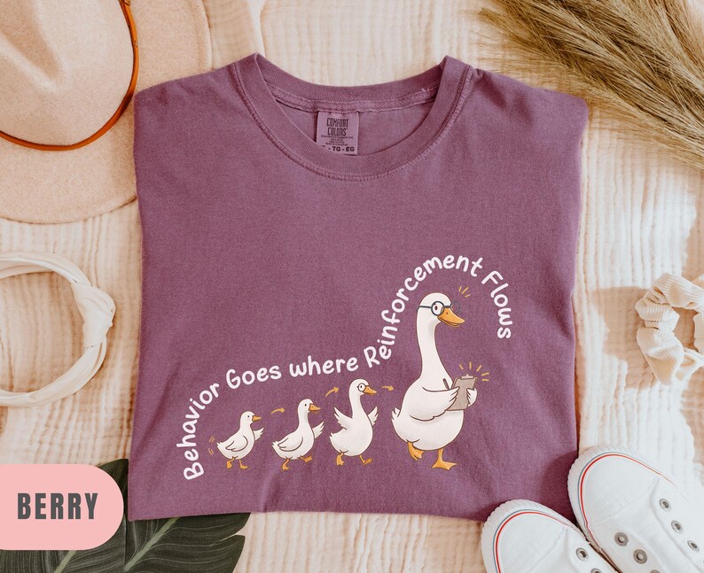 May include: Berry-colored t-shirt with a graphic of a goose wearing glasses and holding a device, followed by three ducklings. The text "Behavior Goes where Reinforcement Flows" is arched around the goose. White sneakers are at the bottom.