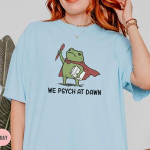 May include: A light blue t-shirt featuring a graphic of a green frog wearing a red cape, holding a red pen and a white book. The text below the frog reads WE PSYCH AT DAWN in black capital letters. The shirt has short sleeves and a crew neckline.