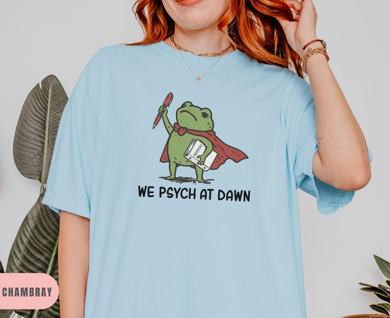May include: A light blue t-shirt featuring a graphic of a green frog wearing a red cape, holding a red pen and a white book. The text below the frog reads WE PSYCH AT DAWN in black capital letters. The shirt has short sleeves and a crew neckline.