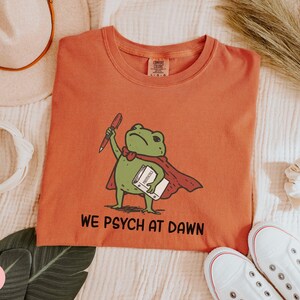 May include: A terracotta-colored t-shirt featuring a graphic of a green frog wearing a red cape, holding a red pen and a white book labeled "PROTOCOLS". Below the frog, the text "WE PSYCH AT DAWN" is printed in black, bold, sans-serif letters.
