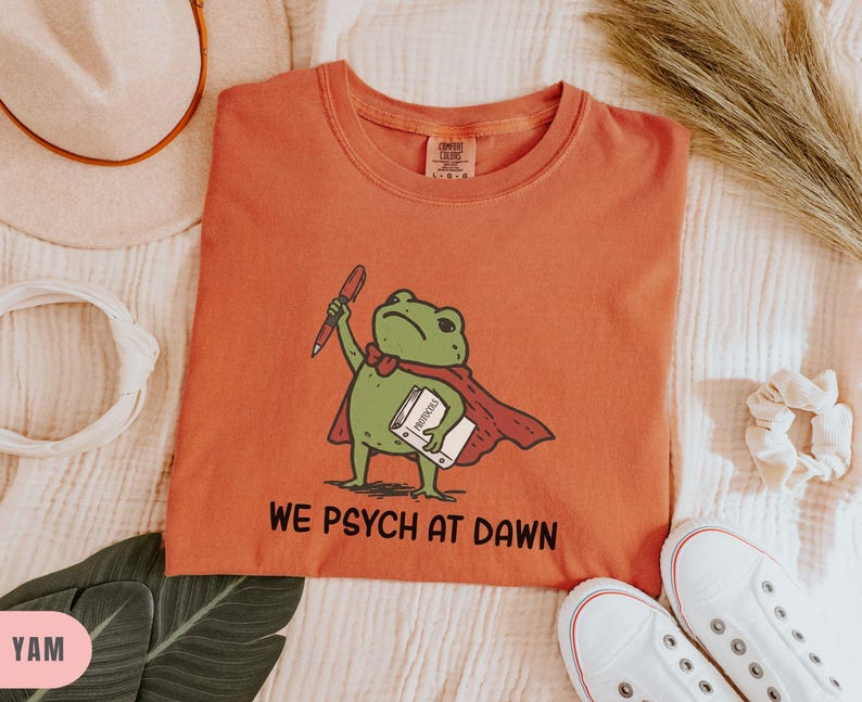 May include: A terracotta-colored t-shirt featuring a graphic of a green frog wearing a red cape, holding a red pen and a white book labeled "PROTOCOLS". Below the frog, the text "WE PSYCH AT DAWN" is printed in black, bold, sans-serif letters.