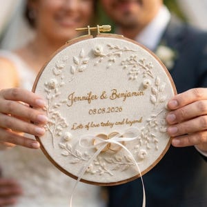 Op de afbeelding: Een crèmekleurige geborduurde ringhouder met de namen "Jennifer & Benjamin" en de datum "08.08.2020" in gouden letters. De houder heeft bloemenborduurwerk en een lint voor de ringen. Een huwelijksherinnering.